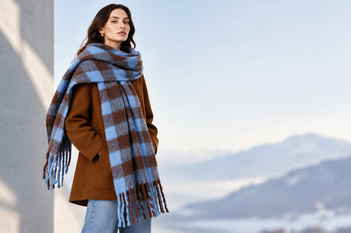 A woman modeling a large plaid scarf in brown and light blue, wrapped around her shoulders over a brown coat and blue jeans, against a plain light gray studio background.